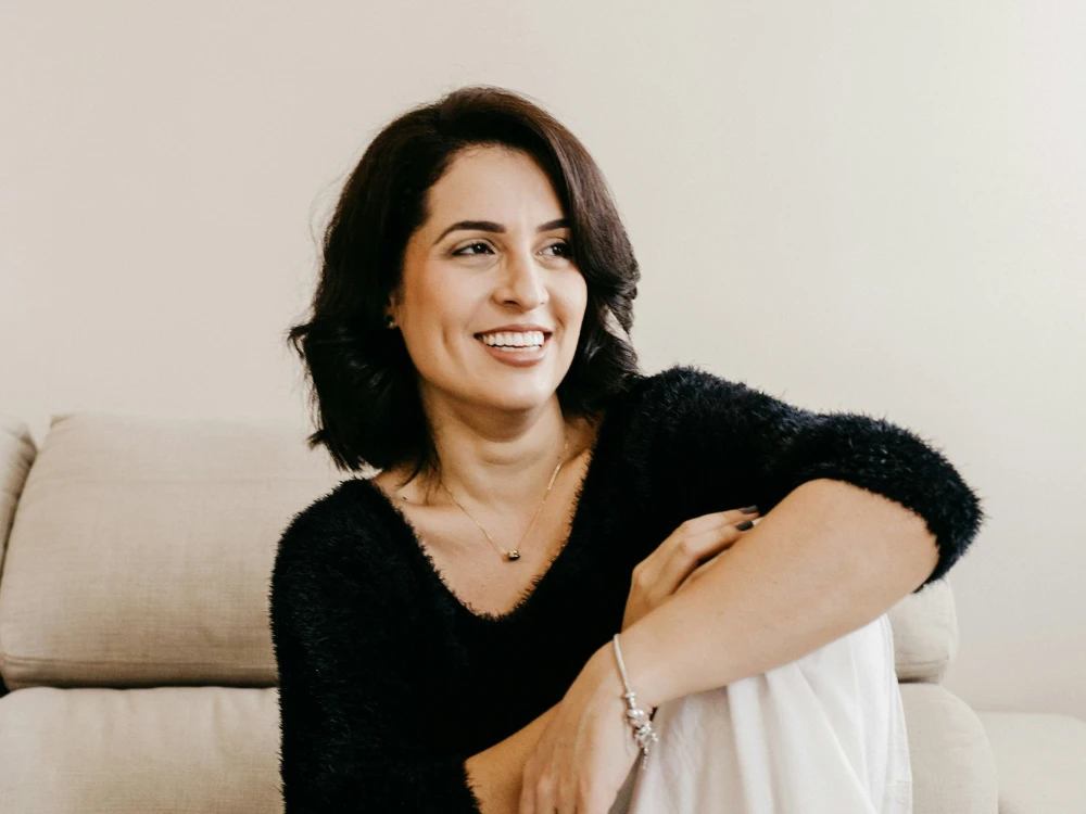 Frau mit dunklen, lockigen Haaren und lächelndem Gesicht, sitzt entspannt auf einem Sofa in einem hellen Raum.