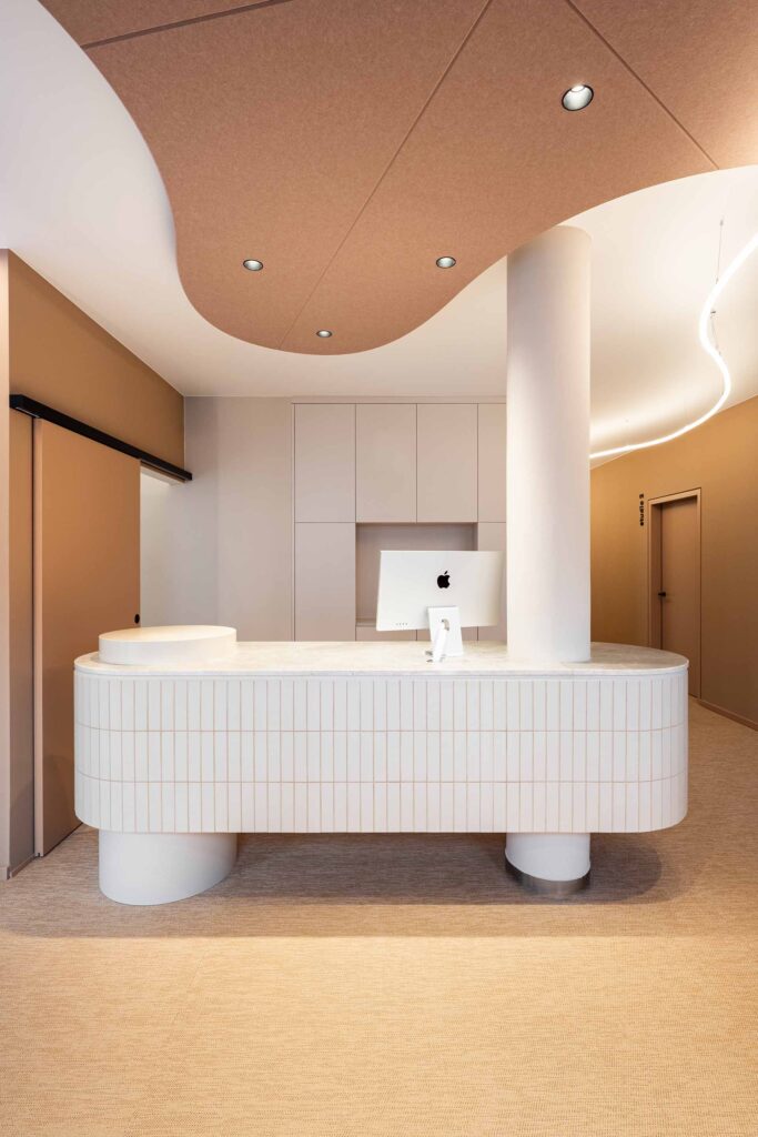 Modernes Empfangs-Interieur im LIV*dental studio: weiße Tresenform, braune Deckenwölbung und eingelassene Spots, mit Apple-Display im Hintergrund.
