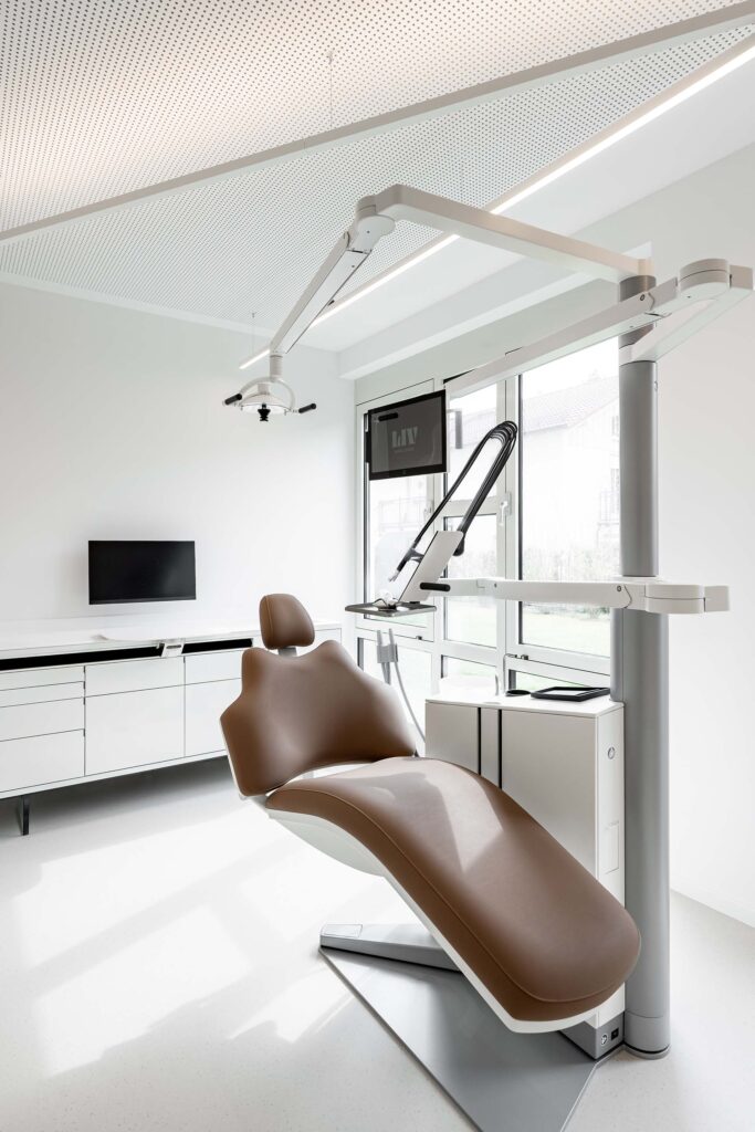 Moderne Behandlungsumgebung im LIV*dental studio: weiße Zahnarzteinheit mit braunem Behandlungssessel, Beleuchtung und Monitor im hellen Raum in Deisenhofen