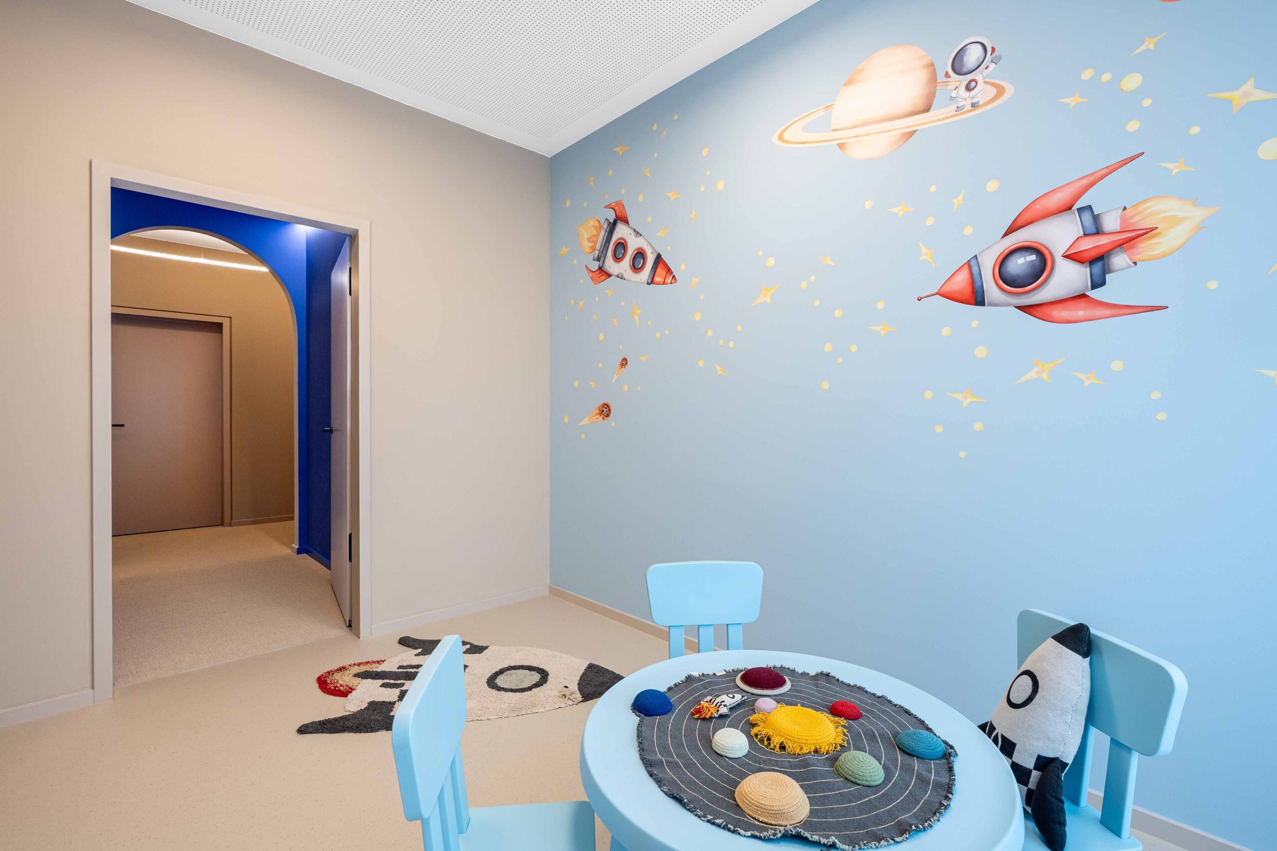 Helles Kinderzimmer im LIV*dental studio mit hellblauer Wand und Raketen-Illustrationen, beigeem Boden und runder Spielstation aus Stoff.