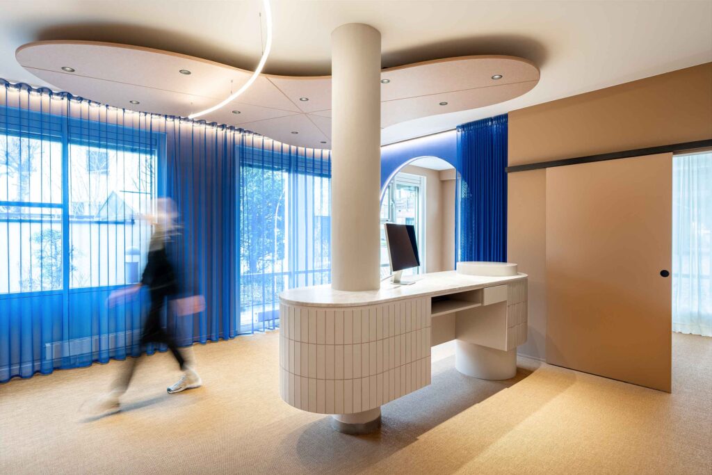 Im hellen Empfangsbereich des LIV*dental studio beim Zahnarzt Deisenhofen mit blauem Vorhang, weißem Empfangstresen und dezentem Kunstlicht.