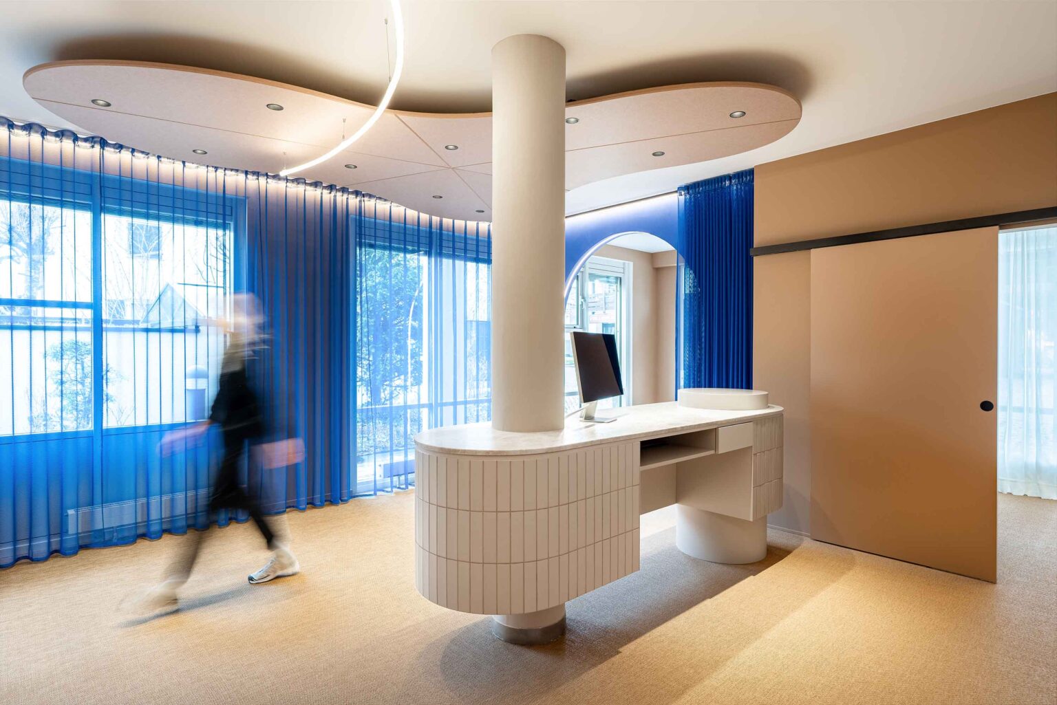 Im hellen Empfangsbereich des LIV*dental studio beim Zahnarzt Deisenhofen mit blauem Vorhang, weißem Empfangstresen und dezentem Kunstlicht.