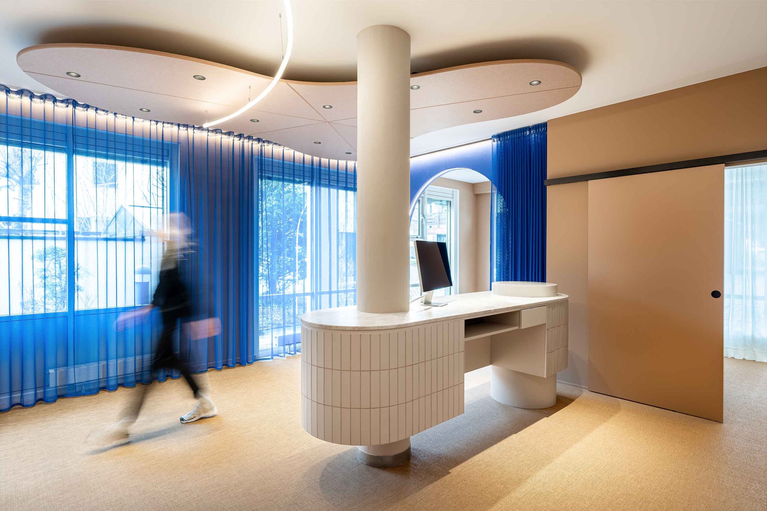 Im hellen Empfangsbereich des LIV*dental studio beim Zahnarzt Deisenhofen mit blauem Vorhang, weißem Empfangstresen und dezentem Kunstlicht.