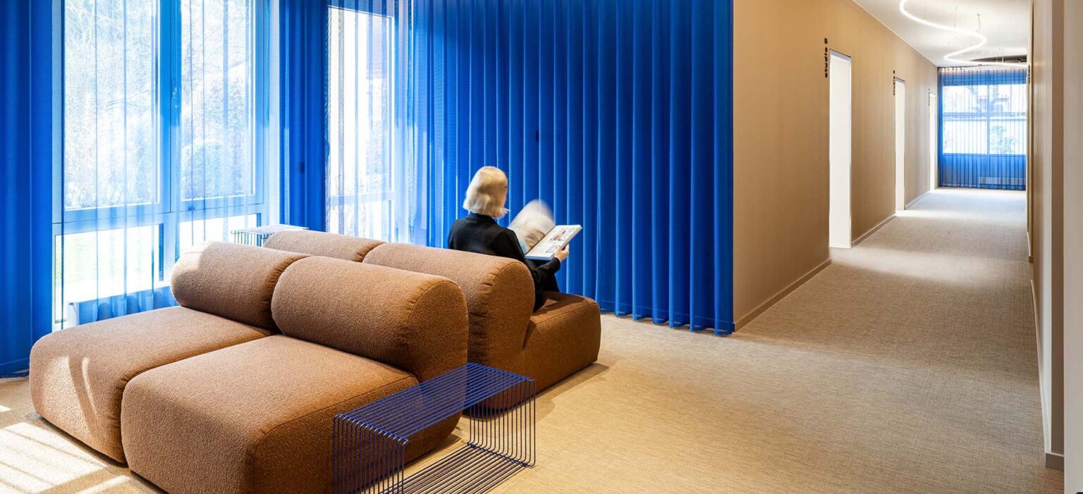 Heller Praxis-Warteraum mit beigen Polstersofas, blauen Vorhängen und modernen, klaren Linien; eine Person liest, große Fenster spenden Licht.