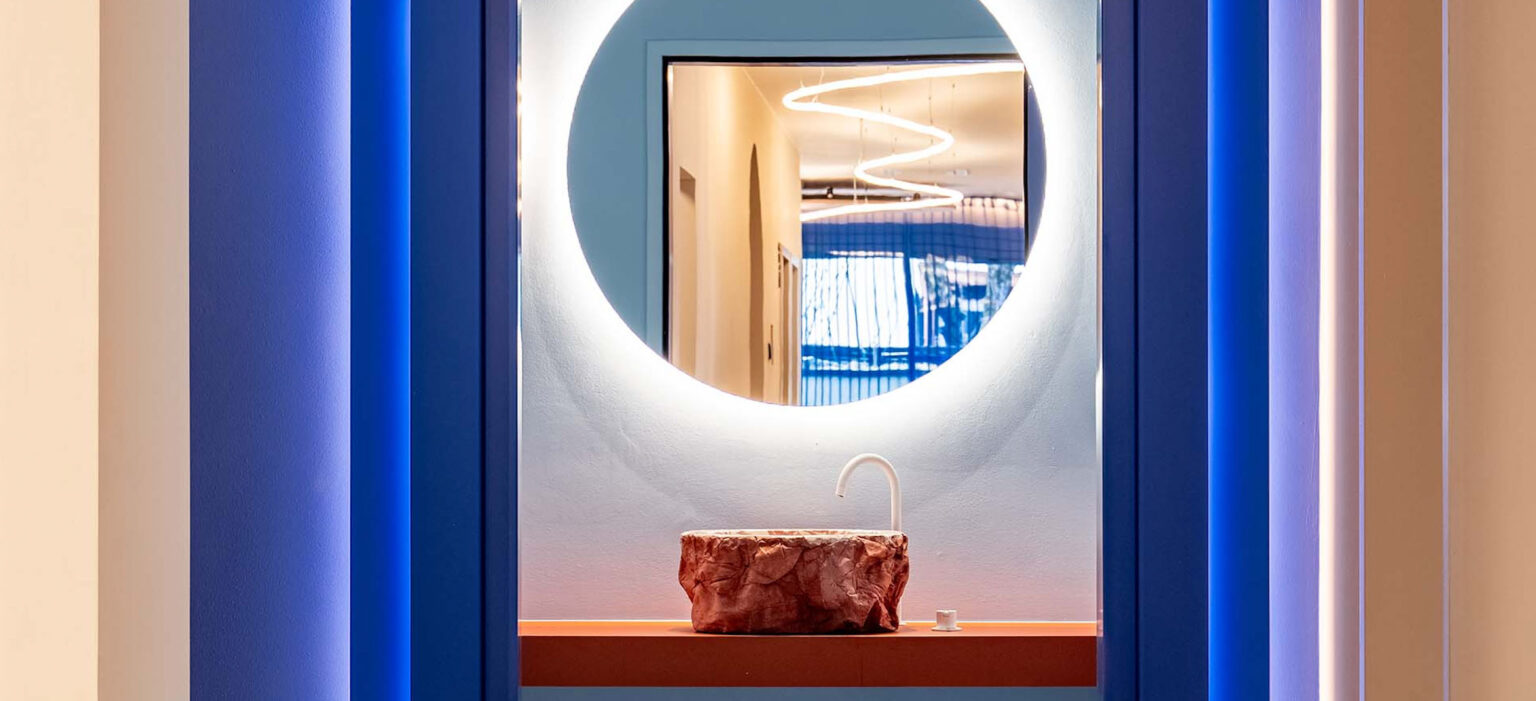 Helle Praxis von LIV*dental studio: ein runder Spiegel über dem Waschtisch, blaues Licht an den Säulen und moderne, klare Linien im lichtdurchfluteten Flur.