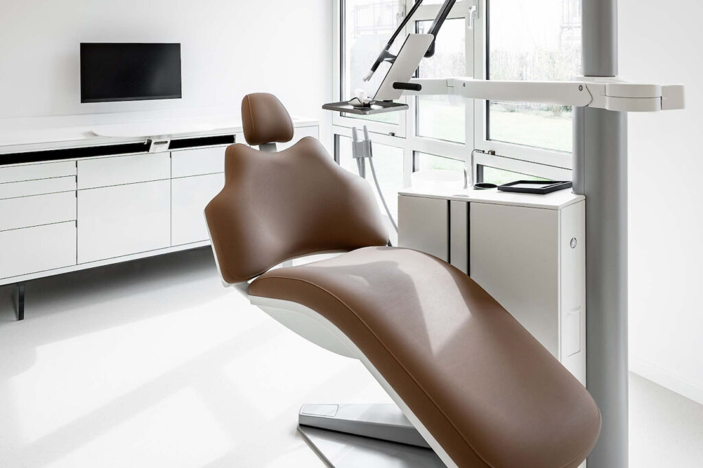 Weißes modernes Behandlungszimmer mit braunem Zahnarztstuhl und klaren Linien der Behandlungseinheit – Präventive Zahnmedizin und Prophylaxe.