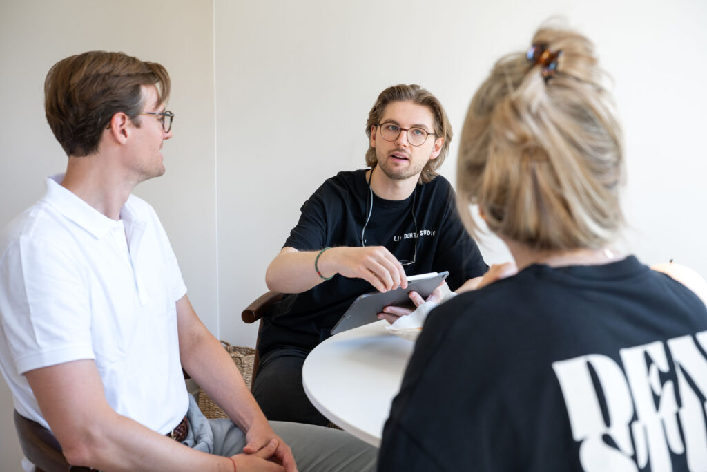 Zwei Zahnärzte beraten eine Patientin am Tisch; einer erklärt etwas und hält dabei ein Tablet in der Hand.