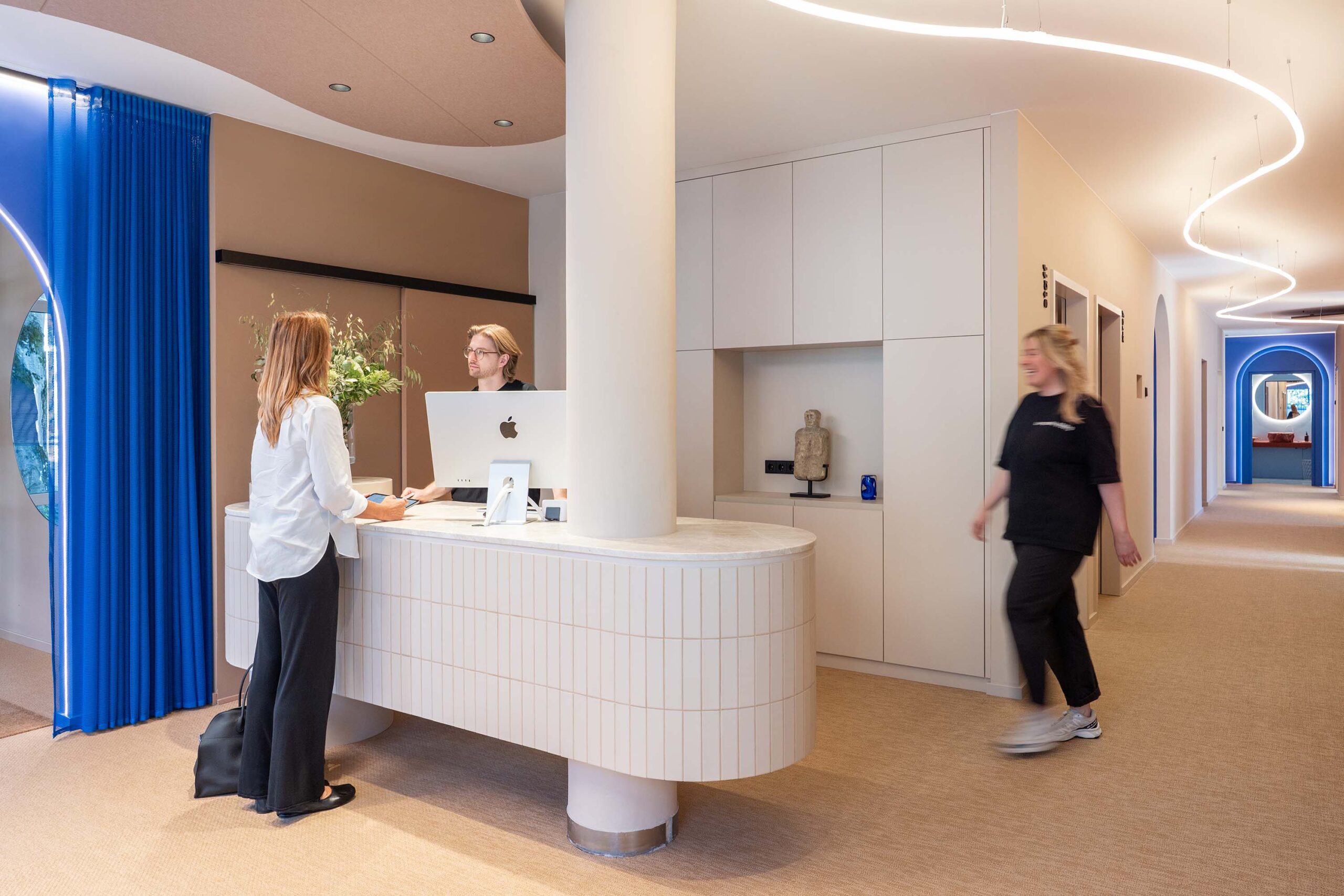 Am Empfang des LIV*dental studio berät ein Mitarbeiter eine Besucherin, während eine Mitarbeiterin durch die helle Zahnarztpraxis geht.