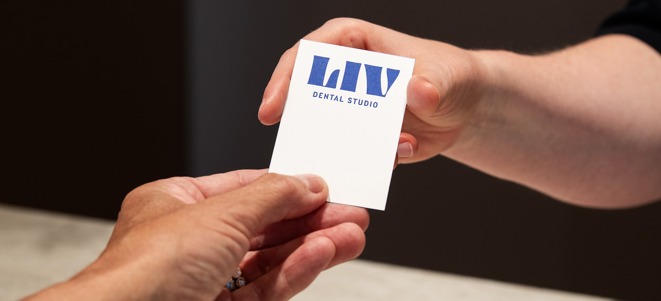 Zwei Hände reichen eine weiße Karte mit blauem Logo und dem Schriftzug „LIV DENTAL STUDIO“ der Zahnarztpraxis in Deisenhofen.