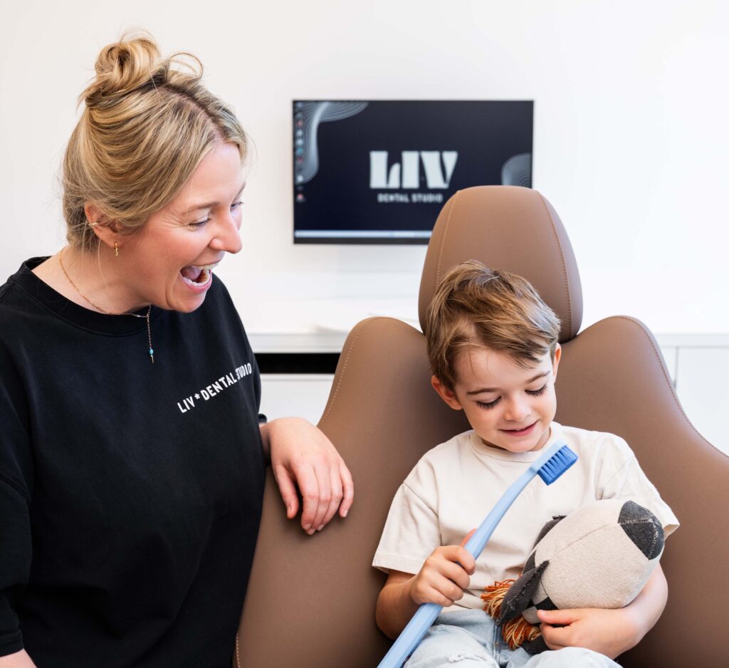 Eine Mitarbeiterin von LIV Dentalstudio übt mit einem Jungen spielerisch Mundbewegungen; er hält eine Zahnbürste und ein Stofftier.