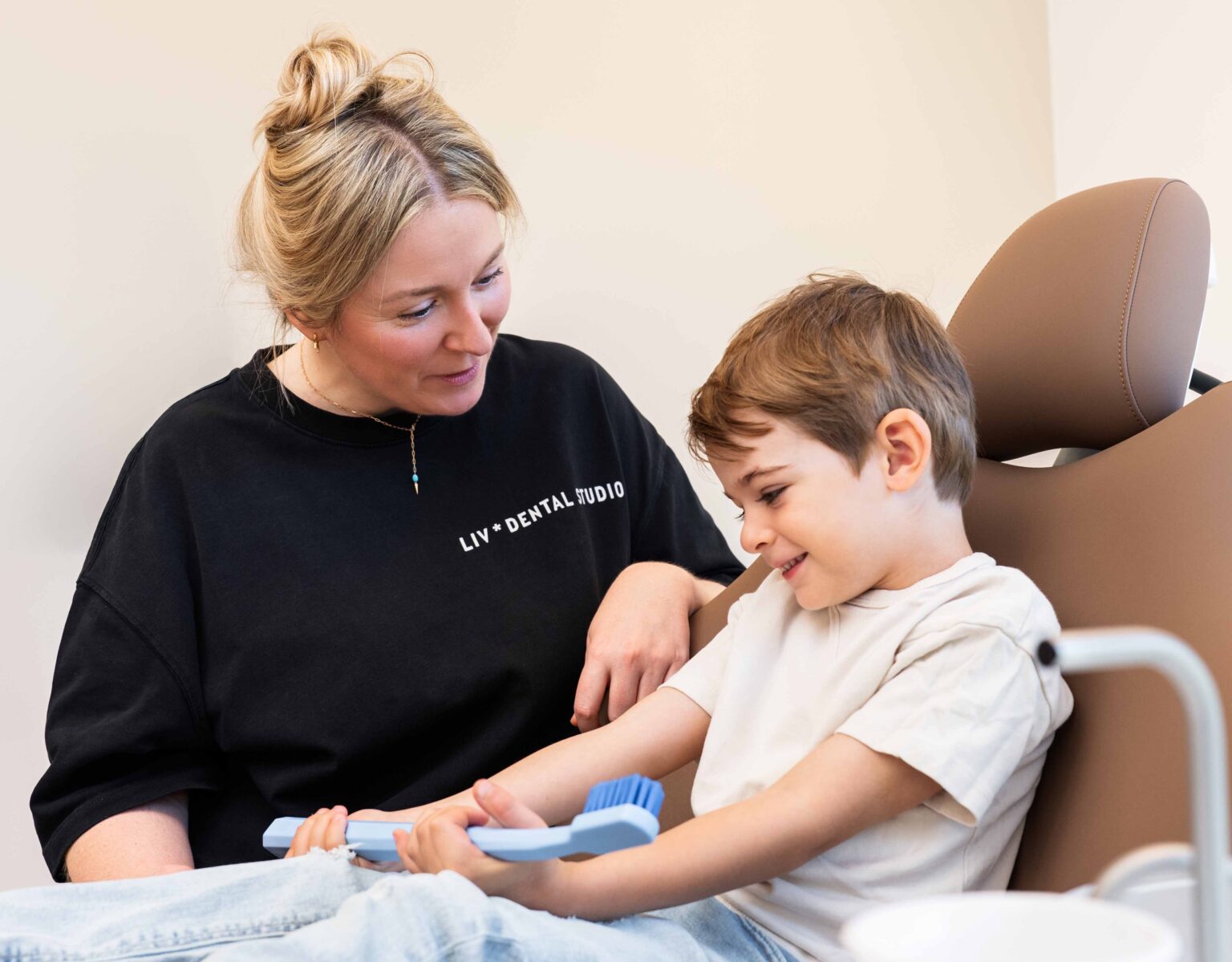 Eine Mitarbeiterin von LIV * dental studio begleitet einen Jungen im Behandlungsstuhl, der lächelnd eine große Zahnbürste hält.