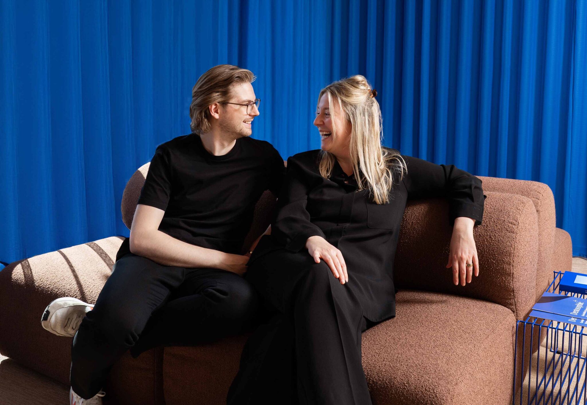 Zwei lächelnde Teammitglieder von LIV dental studio sitzen auf einem Sofa und schauen sich vor blauem Vorhang fröhlich an.