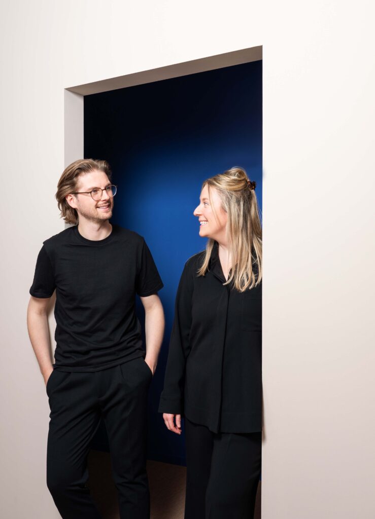 Smiling LIV dental studio team members Laura Bahlmann and Vicco Geßner chat in black clothing beside a deep blue doorway.